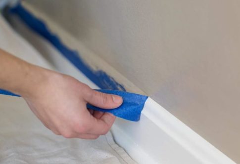A hand pulls painter's tape off a baseboard after painting